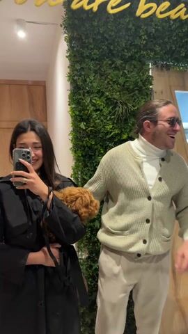 A woman, a man, and a small brown dog are posing for a selfie. The woman is holding the dog, which is nestled against her chest. She is wearing a black coat and has dark hair. The man is standing next to her, wearing a white turtleneck sweater, a light-colored cardigan, and sunglasses. They are both smiling. Behind them, there is a wall covered in greenery and a sign with yellow cursive writing. The setting appears to be indoors, possibly in a shop or lobby.