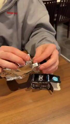 A person wearing a grey sweatshirt sits at a wooden table, carefully unwrapping a small silver foil package. After opening the foil, the person reveals a clear plastic package containing a small, gold-colored, bear-shaped figure. A black and white box is also on the table, bearing a blue symbol resembling an at sign. The person is focused on unwrapping the item. A wooden chair is partially visible in the background.