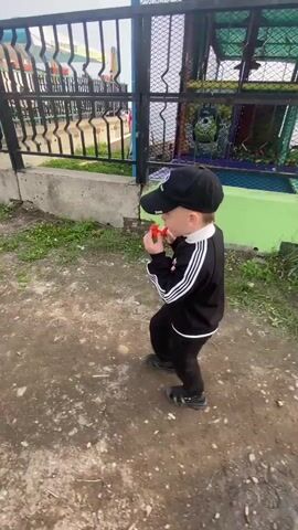A young boy wearing a black baseball cap and a black tracksuit with white stripes on the sleeves walks towards the camera while holding a small red object to his mouth. He is outdoors on a dirt path with patches of grass. Behind him, a black metal fence runs along a concrete wall. Beyond the fence, there are some colorful structures that appear to be part of a playground or amusement area, and in the far distance, there is a body of water. The sky is overcast.