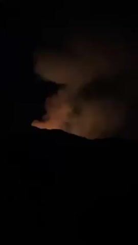 Here is a description of the video:

The video shows a fire burning on a hillside at night. The fire is visible as a patch of orange light through a cloud of smoke. The surrounding area is dark.