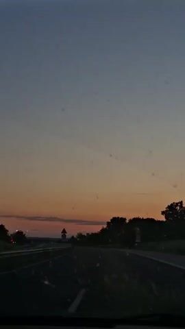 The video shows a car driving on a highway during dusk or dawn. The sky is a gradient of blue and orange. The view is through the car windshield, with trees lining the side of the road. Road signs are visible. Music is playing in the background.