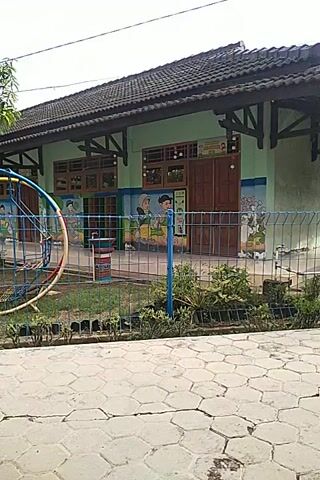 The video shows an outdoor view of a building, likely a school or kindergarten, with a children's playground in the foreground. The building is a single-story structure with a dark roof and light-colored walls, decorated with colorful murals and designs. The playground features various play structures made of metal and plastic, painted in bright colors like blue, red, and yellow. A tree with lush green foliage is visible near the playground, and a wire fence separates the play area from a paved walkway. The camera moves slightly, offering a panning view of the scene from a low angle. The overall atmosphere is bright and cheerful.
