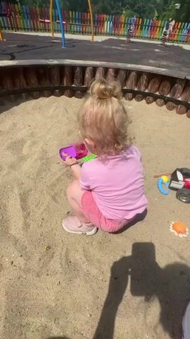 The video begins with a shot of a child playing in a sandbox. The child is wearing a pink shirt and pink shorts and is holding a purple and green toy. The camera then pans to a view of a park with a colorful fence and trees. A young woman is then seen in the foreground, wearing glasses and a tank top. She is sitting on what appears to be roller skates. The video ends with a shot of the woman.