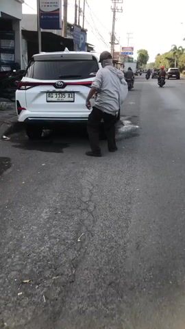 A man wearing a gray hoodie, dark pants, and a light-colored baseball cap is standing by the rear passenger side of a white Toyota Avanza. He is leaning down and appears to be interacting with the car's bumper or rear wheel. The car has an Indonesian license plate that reads "AG 1135 XI". The scene is on an asphalt road with some water or liquid on the ground. In the background, there is a building with signage and a motorcycle rider on the road. The lighting suggests it is daytime.