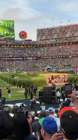 Here’s a description of the video:

The video shows a live performance on the field of a packed stadium. A group of performers are on a pink stage with greenery around it. Other performers are wearing tan clothing and performing on the field. Many people are standing on the field filming. A large screen displays the team logo. The crowd in the stadium is very dense.