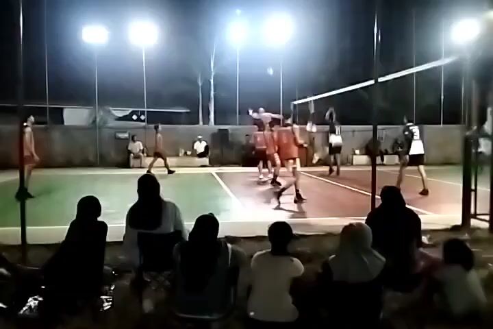 The video shows a volleyball game at night. The court is lit by bright lights. There are players on both sides of the net, and spectators are sitting on the ground watching the game. The players are wearing uniforms, and some of the spectators are wearing headscarves. The game appears to be in progress, with the ball being served or hit. The atmosphere seems casual, with a mix of players and spectators of various ages.