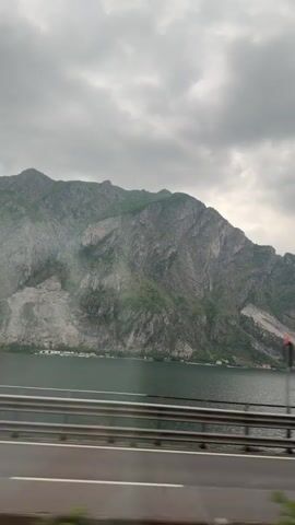 The video shows a landscape of a mountain and lake, seen from a vehicle moving on a road. The mountain is large and rugged, with a mix of green vegetation and exposed rock. The lake is calm and its surface reflects the cloudy sky. The road is lined with a metal guardrail and has yellow painted lines. As the vehicle moves, it passes under the arches of a bridge or overpass. The sky is overcast with grey clouds. No people are visible in the video.
