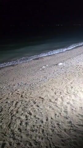 A nighttime view of a sandy beach with the ocean in the background. The sand is light brown and covered in footprints. The waterline is visible with small waves crashing. The ocean is dark, and distant lights can be seen on the horizon. The beach is illuminated by a bright light from the right side of the frame.