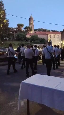A group of people in uniform, holding instruments, are walking in what appears to be a town square. The sky is dark, suggesting it is either dawn or dusk. In the background, there is a building with a tower, possibly a church, and trees. A white tablecloth covers a table in the foreground.