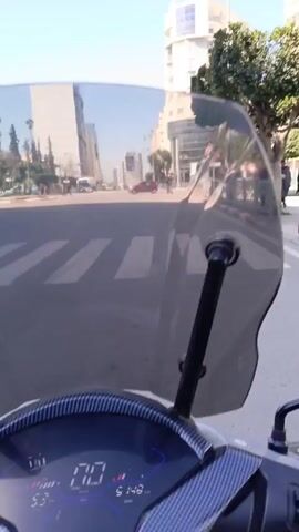 The video shows a street scene in what appears to be a city or town. It's daytime, with a clear blue sky. The camera is mounted on a vehicle, possibly a scooter or motorcycle, giving a first-person perspective of someone driving down the street. There are buildings on either side of the road, including a modern-looking structure with a rounded design. There are crosswalks painted on the street, and cars are visible moving in both directions.  Palm trees and other greenery line the sides of the road. People are seen walking on the sidewalks. The general impression is of a sunny, urban environment.