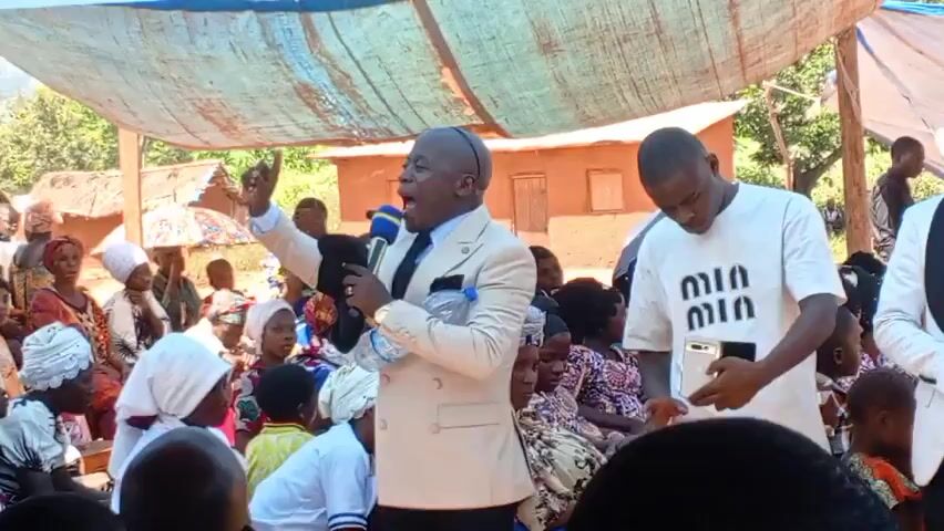 A man in a suit is speaking into a microphone to a seated crowd.  He has a bottle of water in his hand.  Several people are standing behind him. One man behind him is using a cell phone. The event is taking place outdoors under a makeshift canopy. There are houses visible behind the crowd.