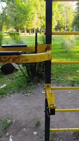 The video captures a walk through an outdoor park, likely in Fastiv, Ukraine, with a focus on a recreational area equipped with exercise machines and a climbing frame. The time of day appears to be late afternoon, as indicated by the warm, golden sunlight filtering through the lush green trees. The weather seems pleasant and dry, with a clear sky and verdant grass suggesting a healthy growing season.

The primary subjects are the various pieces of outdoor gym equipment. A prominent feature is a black and yellow climbing frame with multiple horizontal bars, where a green backpack is hanging from one of the yellow support beams. Further along, the camera pans to show a series of other yellow and black exercise machines, each designed for different physical activities, arranged in a row. These machines are situated on a dirt and gravel path, surrounded by grass and trees.

In the background, the park setting is evident. Trees with full canopies dominate the scene, casting shadows on the ground. A paved pathway winds through the park, suggesting a well-maintained public space. The overall atmosphere is serene and peaceful, conveying a sense of tranquility and a healthy outdoor environment. There are no people visible in the video, and no text is present. The scene suggests a moment of quiet observation in a public park, perhaps before or after a workout.