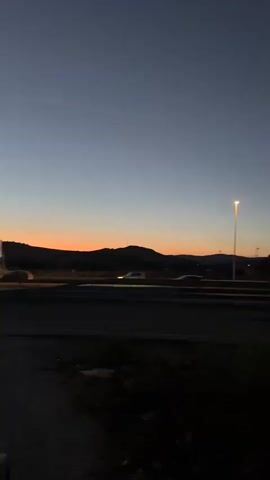 The video shows a twilight scene. In the foreground, a dark, unlit area with some sparse vegetation is visible. A paved road runs horizontally across the midground, with light trails from vehicles indicating movement. To the left of the road, a distant hill is silhouetted against the fading light. A tall streetlamp stands prominently in the center of the midground, emitting a bright light. The sky above transitions from a deep blue at the top to a soft orange and yellow hue near the horizon, suggesting the last moments of sunset. The overall atmosphere is serene and quiet.