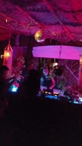 The video shows a nighttime scene at what appears to be a club or bar. Several people are standing in the foreground, their faces mostly obscured in shadow, with a DJ performing on a console behind them. The DJ is a person with short hair, wearing a dark tank top. The setting is dimly lit with pink and purple lights illuminating the scene. A disco ball hangs from the thatched roof, casting reflections. Decorations include plants and a wooden structure resembling a surfboard suspended above the DJ. The atmosphere is festive and energetic, with music playing.