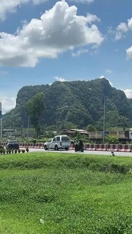 The video shows a road with traffic and a large, forested mountain in the background. The sky is blue with white clouds. The road is divided by a barrier with red and white markings. Several vehicles are visible, including cars, a white SUV, and a motorcycle. A person is riding the motorcycle. In the foreground, there is a field of green grass. The overall scene appears to be a sunny day in a rural or semi-rural area.