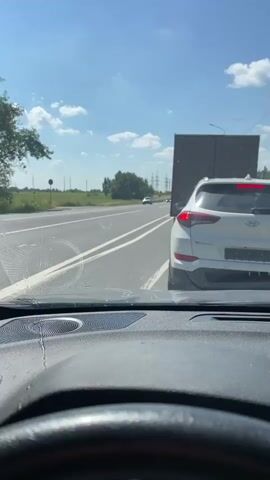 The video shows the interior of a car, with the driver's perspective looking forward. A white SUV is in front, and a large, dark truck trailer is to the right of the SUV. Beyond the vehicles, there is a gas station with pumps and a canopy. A man wearing a black t-shirt and pants is walking away from the SUV towards the gas station. The sky is blue with scattered clouds, and there are trees and fields visible in the distance. A fuzzy, multi-colored charm hangs from the rearview mirror.