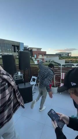 This media file captures a lively rooftop gathering likely in Mukhatskaro, Georgia, during the late afternoon or early evening. The sky is a clear, soft blue, suggesting fair weather. The setting is an outdoor event space with a modern building in the background, featuring large windows and contemporary architecture, possibly a venue or a residential complex.

The primary subjects are groups of people enjoying themselves. In the foreground, a man is energetically dancing with his arms raised, dressed in a patterned shirt and grey pants. Another individual, seen from behind, is also dancing and wearing a plaid shirt. Further into the scene, on a raised stage, a group of performers are actively engaged. One woman in a white outfit is singing into a microphone, accompanied by two men, one in a white t-shirt and camouflage pants, and another in a white shirt and white pants, also holding a microphone.

The event appears to be a musical performance or a party, with large speakers set up, indicating a sound system for the music. The crowd is diverse, with people of various ages and ethnicities. Many are dressed casually, fitting for an outdoor social event. Some individuals are holding phones, likely capturing the moments. The overall mood seems festive and celebratory, with people dancing, singing along, and interacting with each other.

Notable details include the modern building with some floral decorations visible on its facade. The presence of white folding chairs suggests seating arrangements might be available. The time of day is indicated by the soft lighting and the clear sky. There is visible text on the building in the background, reading "MUAHTSKARO EVENT HALL".