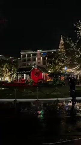 The video shows a festive, dimly lit outdoor scene at night, likely during the Christmas season. In the center is a small, brightly illuminated red building labeled "Santa's House" with a wreath on the front. It is surrounded by smaller Christmas trees and red poinsettias. A tall Christmas tree decorated with lights stands to the right. Buildings are visible in the background with lit windows. String lights are hung in the trees. In the foreground, the ground appears wet and reflective, possibly due to rain, and a person with an umbrella walks across the frame, passing in front of the Christmas decorations. The scene is enclosed by a black rope fence.