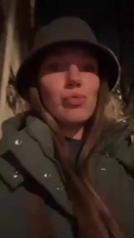 The video depicts a woman walking down a street at night. The woman is wearing a green jacket and a gray bucket hat. She has long blonde hair. The background shows street lights, buildings, and cars. The video is taken from a close-up perspective, focusing on the woman's face. She smiles slightly at the camera.