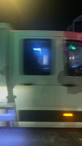 The video shows a street scene at night. A delivery truck passes by in the beginning of the video. A bright blue sign illuminates a small shop across the street. There is a red stoplight, a crosswalk, and a scooter parked in front of the shop. Later, a black car drives past in front of the camera. There is also a black fence on the right side of the street.