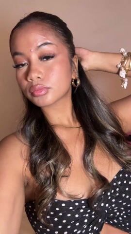 Here is a detailed description of the video:

A young woman is in a close-up selfie video. She has long, wavy brown hair with blonde highlights pulled back at the front. She is wearing gold hoop earrings, a gold necklace, and several bracelets on her right wrist. She is wearing a black and white polka dot strapless top. She looks directly at the camera, flips her hair, sticks her tongue out briefly, and makes a kissy face. The background is a beige wall. The music in the video is upbeat.