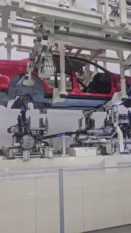 A car chassis is suspended from a robotic assembly line. The chassis is red and appears to be partially assembled. Below the chassis, robotic arms are attaching components to the underside of the vehicle. The scene is well-lit and appears to be a factory or manufacturing facility. The assembly line is moving, as evidenced by the slight blur in the video.