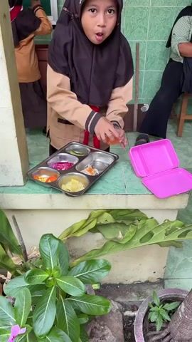 A group of children in school uniforms are gathered outdoors, some interacting with food trays and containers. A young girl in a brown uniform and black hijab is holding a small container of orange liquid over a metal tray with compartments containing various food items. Next to her is an open pink plastic lunchbox. Another person, wearing a green shirt and black hijab, is seated in the background. Further away, two other children in similar uniforms are interacting with food items on trays spread on the floor. The environment appears to be an outdoor area with tiled walls and a concrete floor, with plants in pots visible. The children are engaged in eating or preparing to eat.