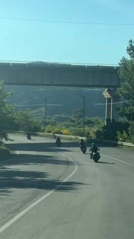 A group of four motorcycles are riding on a winding road under a bridge. The motorcycles are spaced out, with the lead motorcycle farthest away and the last motorcycle closest to the camera. The road curves to the right. The environment is hilly and green, with trees and mountains in the background. The sky is clear and blue. The video appears to be filmed from another moving vehicle.