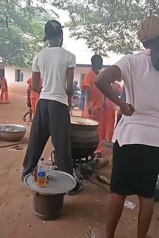 The video shows a group of people in an outdoor setting. In the foreground, a person wearing a white t-shirt and black pants stands facing a large pot over a fire. Next to them, a person in a pink shirt and black skirt stands with their hands on their hips. In the background, several people in orange uniforms are visible near a building. The ground is dirt and there are trees and foliage in the background. The scene appears to be a cooking area, possibly outdoors at an institution.
