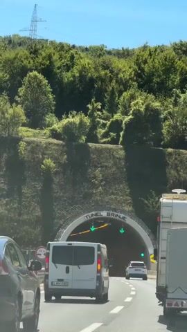 The video shows a view from a car driving towards the entrance of the Brinje Tunnel. The tunnel entrance is visible in the distance, surrounded by green trees and vegetation. The tunnel is labeled "TUNEL BRINJE" above the entrance. Traffic lights, displaying green arrows, are positioned above the tunnel entrance, indicating that it is open. Several vehicles are in front of the camera, including a white van and a dark-colored SUV. The sky is clear and blue.