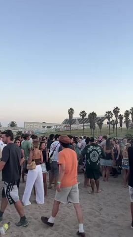 A crowd of young adults gathers on a sandy beach at dusk, illuminated by the warm glow of the setting sun. Some individuals walk around, while others stand in groups, socializing and enjoying beverages. In the distance, a stage with red lights is visible, suggesting a concert or festival. Palm trees line the horizon, and a structure with "SRAM" written in graffiti can be seen in the background. The sky is a clear, light blue, with a hint of orange near the horizon.