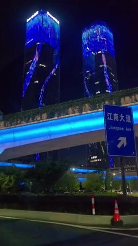 The video displays a vibrant urban night scene, likely in Binjiang District, China. Two tall, modern skyscrapers are prominently featured in the background, their facades illuminated by dynamic LED lights that transition through colors such as blue, purple, and red, creating changing patterns. An elevated road or bridge cuts across the middle ground, with its underside bathed in a striking blue light. Below this structure, green foliage and trees are visible, adding a touch of nature to the urban landscape. In the foreground, a road or street is present, and on the right side, two red and white traffic cones are visible. A blue road sign with white text is also on the right, displaying "江南大道" (Jiangnan Avenue) in Chinese and "Jiangnan Ave" in English, accompanied by a right-pointing arrow. The dark sky confirms it is nighttime.