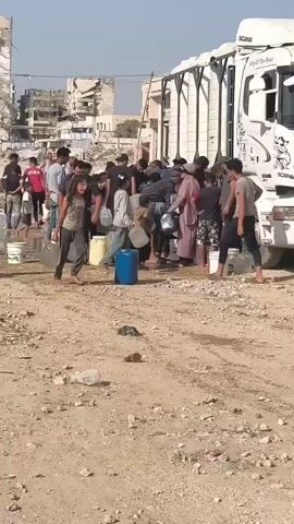 A group of people, including men, women, and children, are gathered around a large white truck, some holding buckets and plastic containers. They appear to be collecting water. The environment is dusty and appears to be an urban area with damaged buildings in the background. Some people are carrying full containers, while others are waiting in line. The truck has some text and graphics on its side. One man in the foreground is walking towards the camera carrying two black buckets. The sky is clear and sunny.