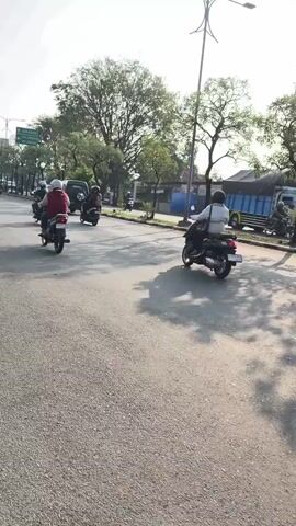 Two motorcycles and a white car are traveling down a paved road on a sunny day. The motorcyclists are wearing helmets. The motorcycle in the foreground has a rider wearing a black jacket and an orange and yellow helmet. The motorcycle behind it has a rider wearing a black jacket and a black helmet. The white car is in front of the second motorcycle and has tinted windows. Trees line the side of the road, and a light pole with what appears to be a cell tower behind it is visible in the background. The scene appears to be in a city or suburban area.