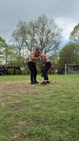 Two men, bare-chested and wearing dark pants, are wrestling in a grassy field. They are locked in a standing grapple, each attempting to gain an advantage. An older man in a vest and trousers stands nearby, seemingly observing or officiating the match. A small crowd of people watches from the edge of the field, with trees and a soccer goal visible in the background. The sky is overcast.