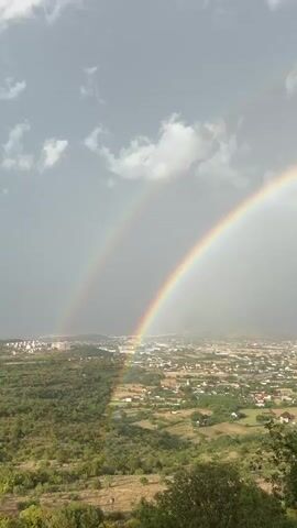 The video shows a landscape with a double rainbow stretching across a cloudy sky. The view is from an elevated position overlooking a town or village nestled in a valley. The foreground consists of dry, brush-covered hills with some green trees. The town below has many small buildings with red roofs, interspersed with patches of green vegetation. The sky is a mix of grey clouds and lighter areas, with the double rainbow prominently displayed. The camera appears to be panning slowly, offering a panoramic view of the scene. There are no people or explicit actions depicted, the focus is on the natural phenomenon of the rainbow and the surrounding environment.