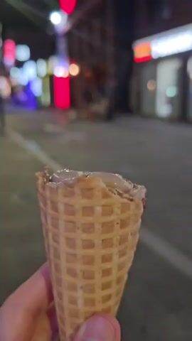 A close-up shot of a person's hand holding a waffle cone with melting ice cream. The background is blurred, showing a street at night with neon lights and buildings. A person walks by in the background, blurred. The scene appears to be a busy urban area.