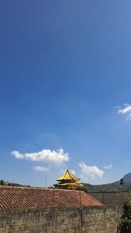 A vertical video pans across a landscape. In the foreground, a stone wall runs horizontally, topped by red-tiled roofs of houses and some greenery. Behind the wall, a distinctive yellow-roofed building, possibly a temple or pagoda, is visible. Further in the background, a large, misty mountain range dominates the horizon. The sky is a clear, bright blue with a few scattered white clouds. The camera movement is smooth, suggesting a deliberate panning motion to capture the scene. No people or animals are visible. The overall impression is one of a serene, natural landscape with architectural elements.