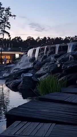 The video shows a man-made waterfall cascading over rocks into a pond. A wooden walkway leads towards the pond. The scene is set at dusk, with trees silhouetted against a twilight sky. The lighting suggests it is an outdoor setting at a park or resort. There is one person visible on the left side of the frame, walking towards a building. The water appears to be flowing from a structure at the top of the rocks. The overall mood is serene and tranquil.
