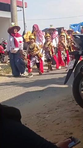 This video shows a group of children in traditional costumes walking down a dirt road. The children are wearing elaborate gold headpieces and colorful outfits with red sashes. They appear to be part of a procession or parade. Adults are walking alongside them, some also in traditional attire, including headscarves. A motorcycle is parked on the right side of the frame. The environment appears to be outdoors in a sunny, possibly rural setting, with buildings and tents visible in the background. The camera is positioned low, looking up at the participants.