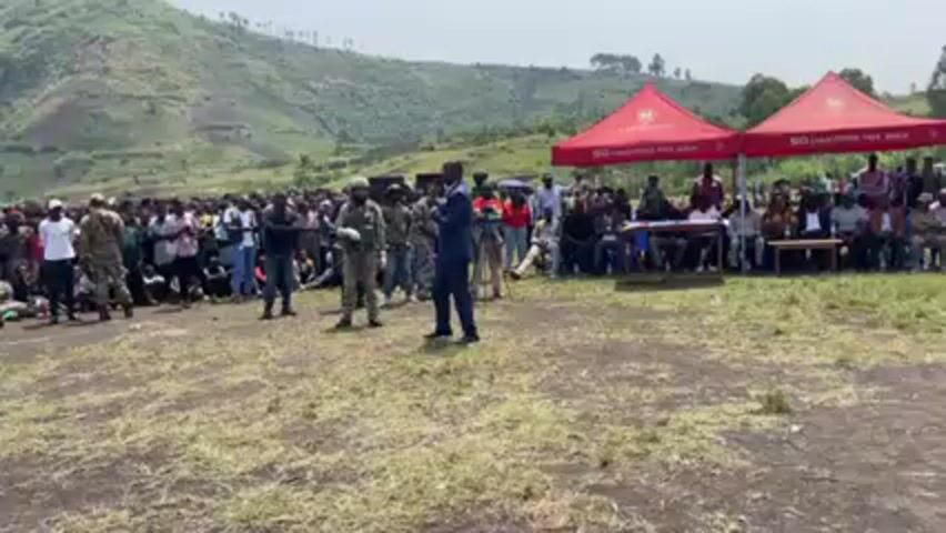 A man in a suit is speaking to a large crowd gathered in an open field against a backdrop of lush green hills. He is surrounded by several people in military uniforms. The crowd appears to be a mix of men, women, and children, some sitting on the ground and others standing. Two red canopies are set up to the right, with people seated at tables beneath them. A cameraman is filming the speaker. The atmosphere seems to be that of an official event or address.