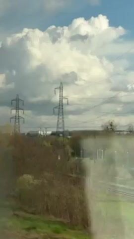 The video shows a view from a moving train. Power lines and towers are visible in the distance, set against a cloudy sky. The train tracks are seen curving through the landscape. Vegetation, including trees with sparse foliage, is also present. The view is partially obscured by reflections on the train window.