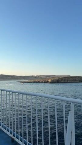 The video shows a view from a moving ferry on a clear, sunny day. The water is a deep blue, and the sky is a bright, cloudless blue. In the distance, there is a rocky coastline with some buildings on top. The ferry has a white railing in the foreground, with some blue paint visible at the bottom left of the frame. The camera is panning slightly to the right, following the coastline.