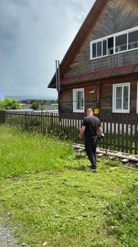 A man in a black t-shirt and black pants is using a string trimmer to cut grass in front of a wooden house. The house has a dark red roof and white framed windows. A wooden fence runs along the side of the property, separating the lawn from a grassy area with wildflowers. The sky is cloudy.