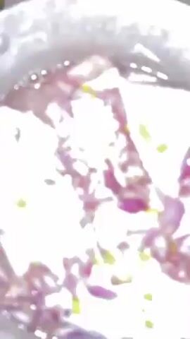 A top-down, static shot shows raw poultry meat in a stainless steel pot filled with water, along with chopped red onions and small pieces of yellow seasoning. The pot is positioned on a tan countertop. A brief voiceover is heard.