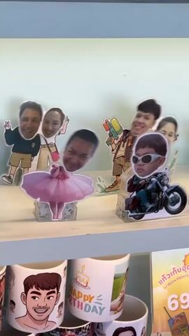 The video shows a close-up, panning shot of a shelf displaying various novelty items. The items are primarily cardboard cutouts of people with exaggerated faces, placed on clear plastic stands. Some cutouts are depicted in action poses, such as riding a motorcycle, holding water guns, or dressed in a ballerina outfit. In the background, blurred cutouts of other people are visible. Below the shelf, several ceramic mugs are displayed. Some mugs feature cartoonish portraits of men and women, while one mug is decorated with a "Happy Birthday" design and a cake graphic. A sign with Thai text and the number "690B" is also visible, suggesting a price. The overall environment appears to be a retail display, likely in a gift shop or novelty store.