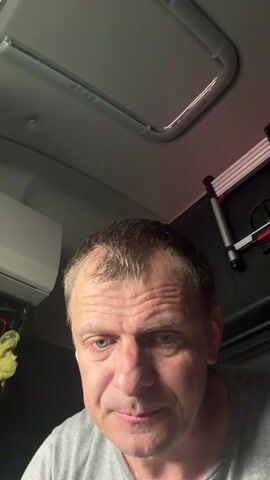 A man is looking down and then up at the camera. The man is wearing a gray t-shirt. The man is in a vehicle. There is a skylight in the ceiling of the vehicle. There is an air conditioner unit on the left side of the image. There are some metal bars on the right side of the image.