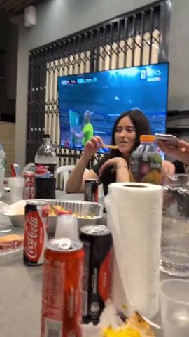 The video shows a group of young adults gathered around a table, appearing to be watching a sporting event on a large television screen in the background. The table is cluttered with food containers, drinks, and paper towels. One person is holding a cell phone, possibly recording or taking a picture. Another person is seen eating. The overall atmosphere seems casual and social.