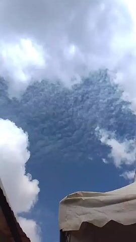 The video shows a partly cloudy sky with a patch of blue visible. There are puffy white clouds on the left and top of the frame, and a field of smaller, patterned clouds in the center. Parts of a roof and a white tent are visible in the lower part of the frame. Music can be heard in the background.