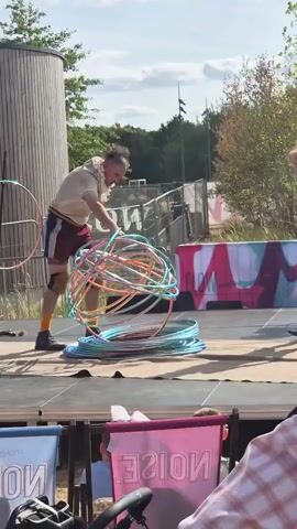 A man is on a stage with a pile of hula hoops. He is wearing shorts, a shirt, and knee pads. He is reaching down and picking up a hula hoop. There is a wooden building behind him and trees in the background. There are people in the foreground sitting in chairs.
